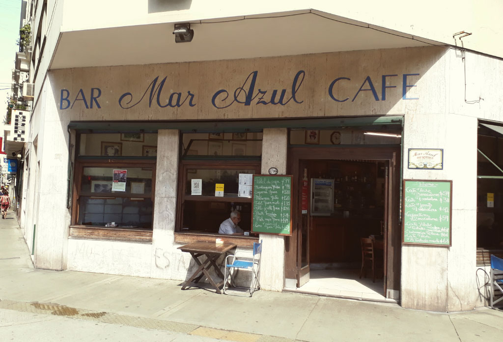 Façade du remarquable bar Mar Azul avec la porte d'entrée ouverte, deux tableaux noirs sur les côtés et deux fenêtres. Par une fenêtre, on peut voir un homme boire du café.
