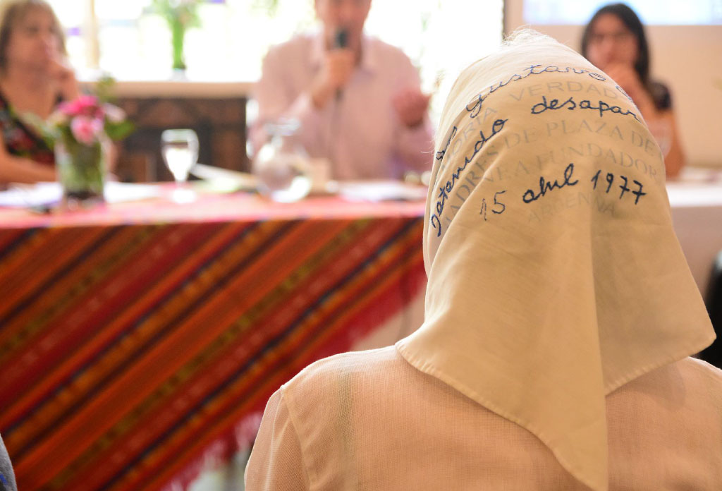 femme dos à une table avec d’autres personnes. La femme porte un foulard blanc autour de sa tête avec l’inscription des données d’un parent disparu.