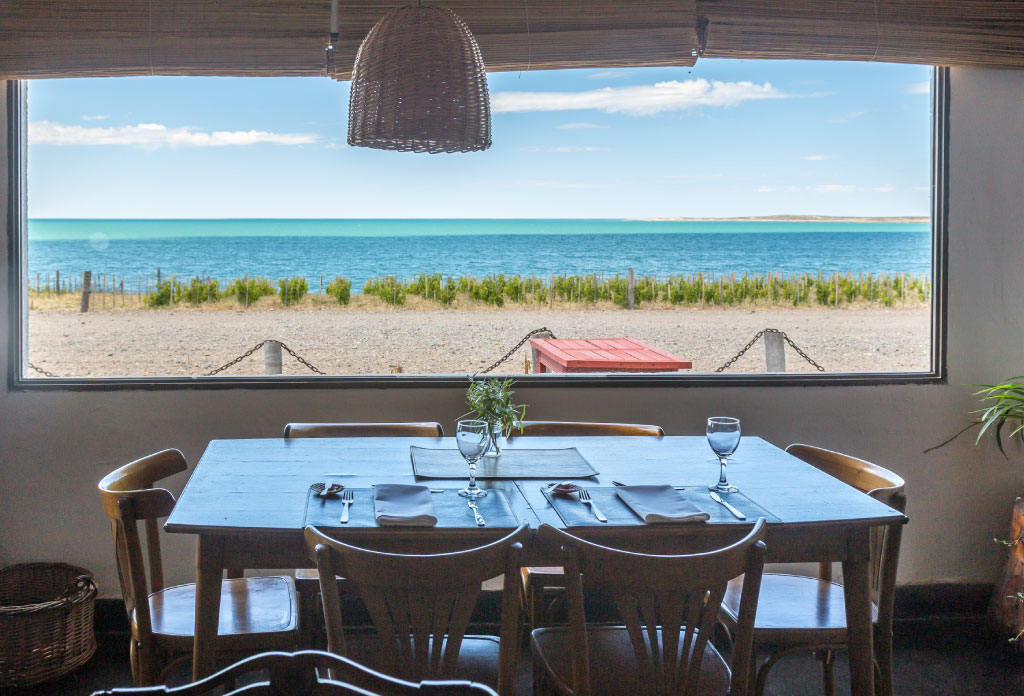 une table, des chaisses et des couverts avec une grande fenêtre dans le restaurante de Bahía Bustamante Lodge