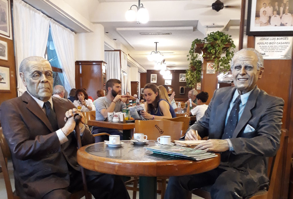 Sculptures des écrivains Jorge Luis Borges et Adolfo Bioy Casares dans le bar La Biela. Derrière se trouvent deux touristes qui mangent, boivent et lisent un livre.