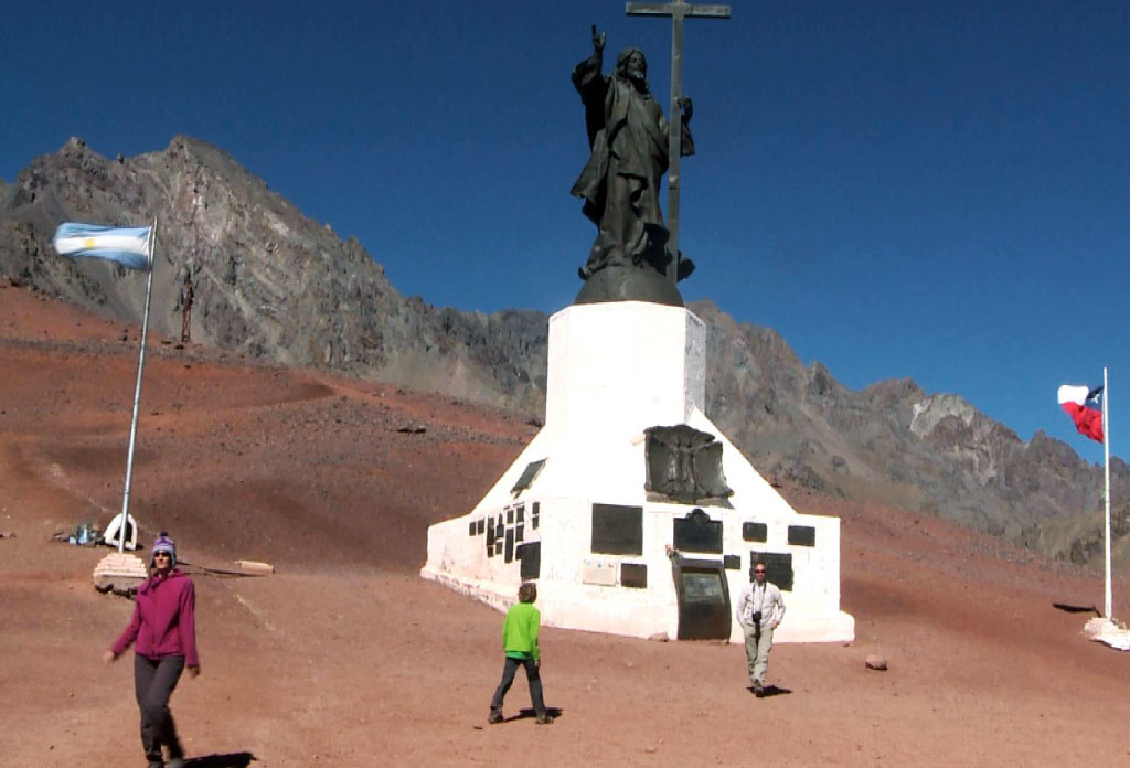 Grande sculpture du christ rédempteur sur un piédestal blanc. derrière on voit des montagnes. Sur ses côtés il y a des gens et des drapeaux de l’Argentine et du Chili.