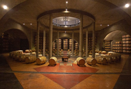 grande salle avec de nombreux tonneaux en bois. Au centre, il y a un piano et autour de lui des colonnes qui soutiennent le plafond. C’est l’intérieur de la cave Salentein.