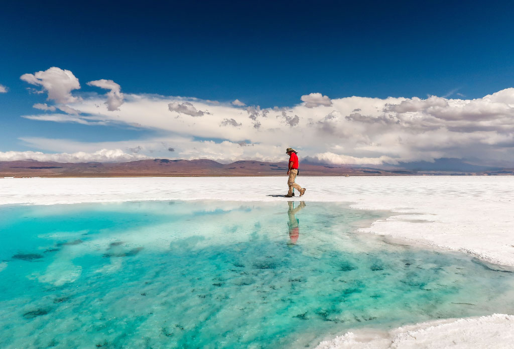 une personne marche sur un plat de sel. On peut voir des montagnes derrière. À côté de lui se trouve un miroir d'eau.
