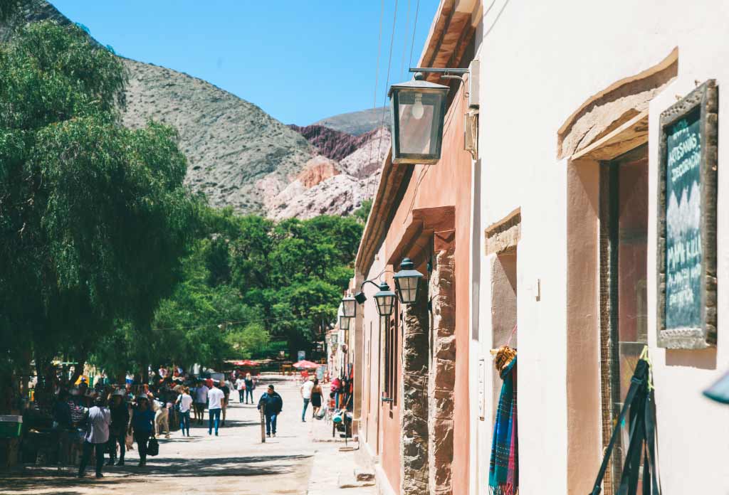 ville de Purmamarca. Une rue avec des gens qui vendent de l'artisanat et devant eux plusieurs magasins avec des murs en adobe et derrière eux la montagne Cerro de los 7 colores.