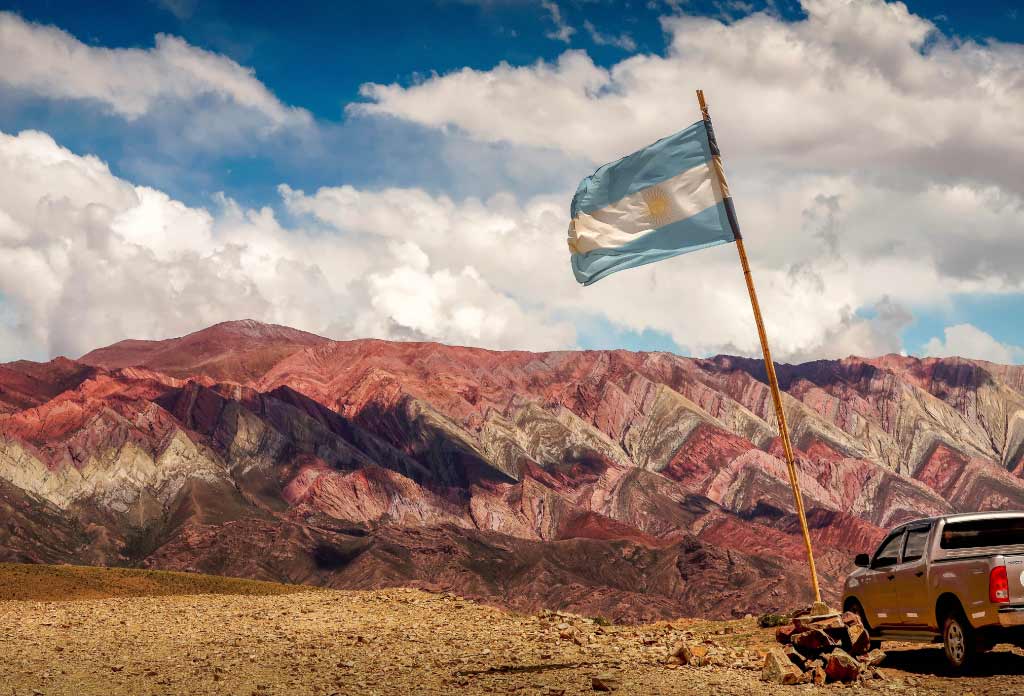Montagne avec beaucoup de couleurs de terre. Devant, il y a une camionnette et un mât avec le drapeau argentin.