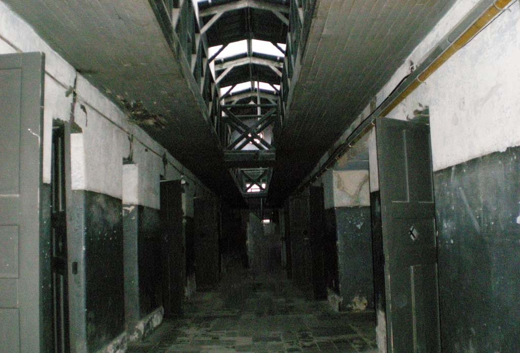 Couloir de l'ancienne prison en mauvais état avec des cellules de chaque côté du couloir.